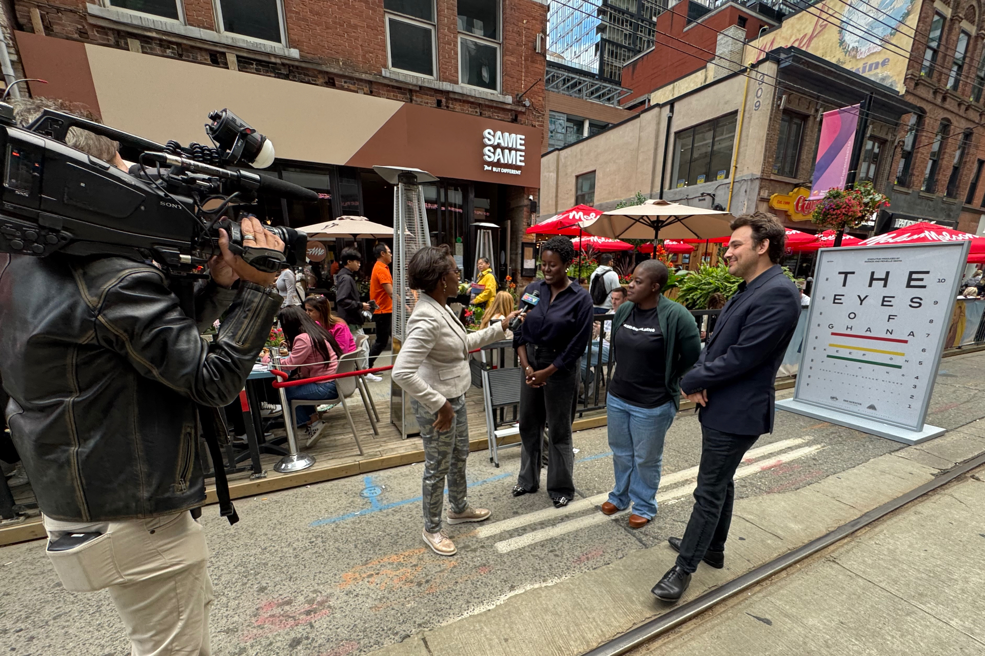 CTV News crew interviews The Eyes of Ghana filmmakers on a Toronto street next to the film's eye-chart poster during the festival premiere.