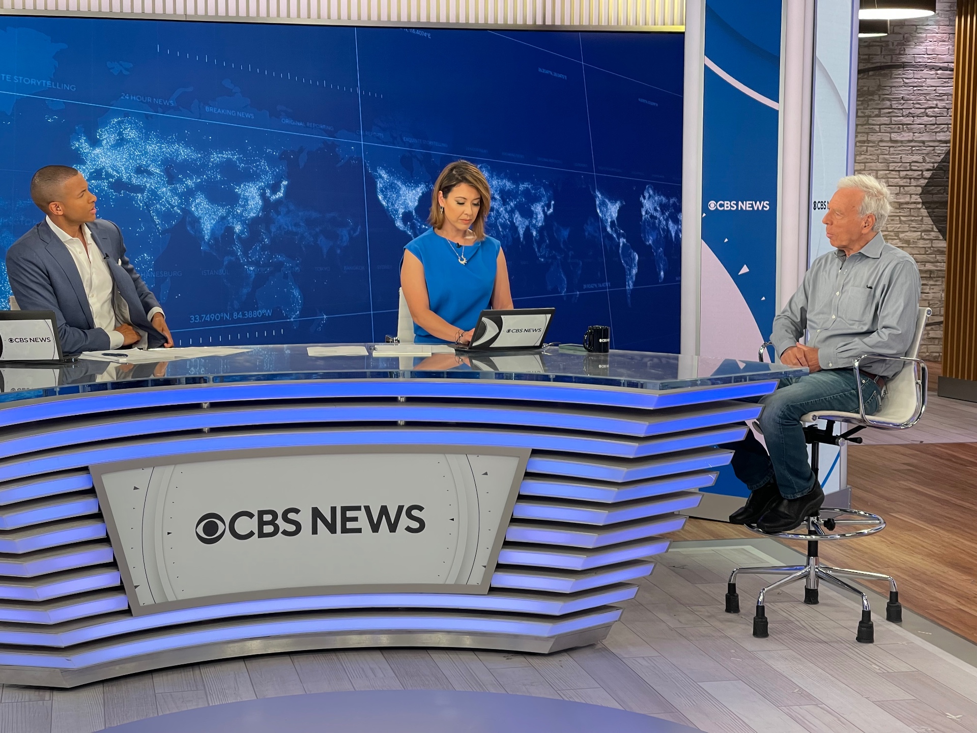 Director sits across from CBS News co-anchors Errol Barnett and Lana Zak for a live interview at the anchor desk.