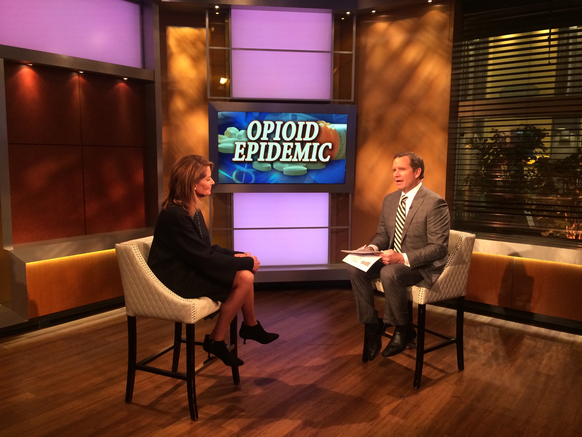 Client interviewed by a news anchor on set beneath an 'Opioid Epidemic' broadcast graphic.