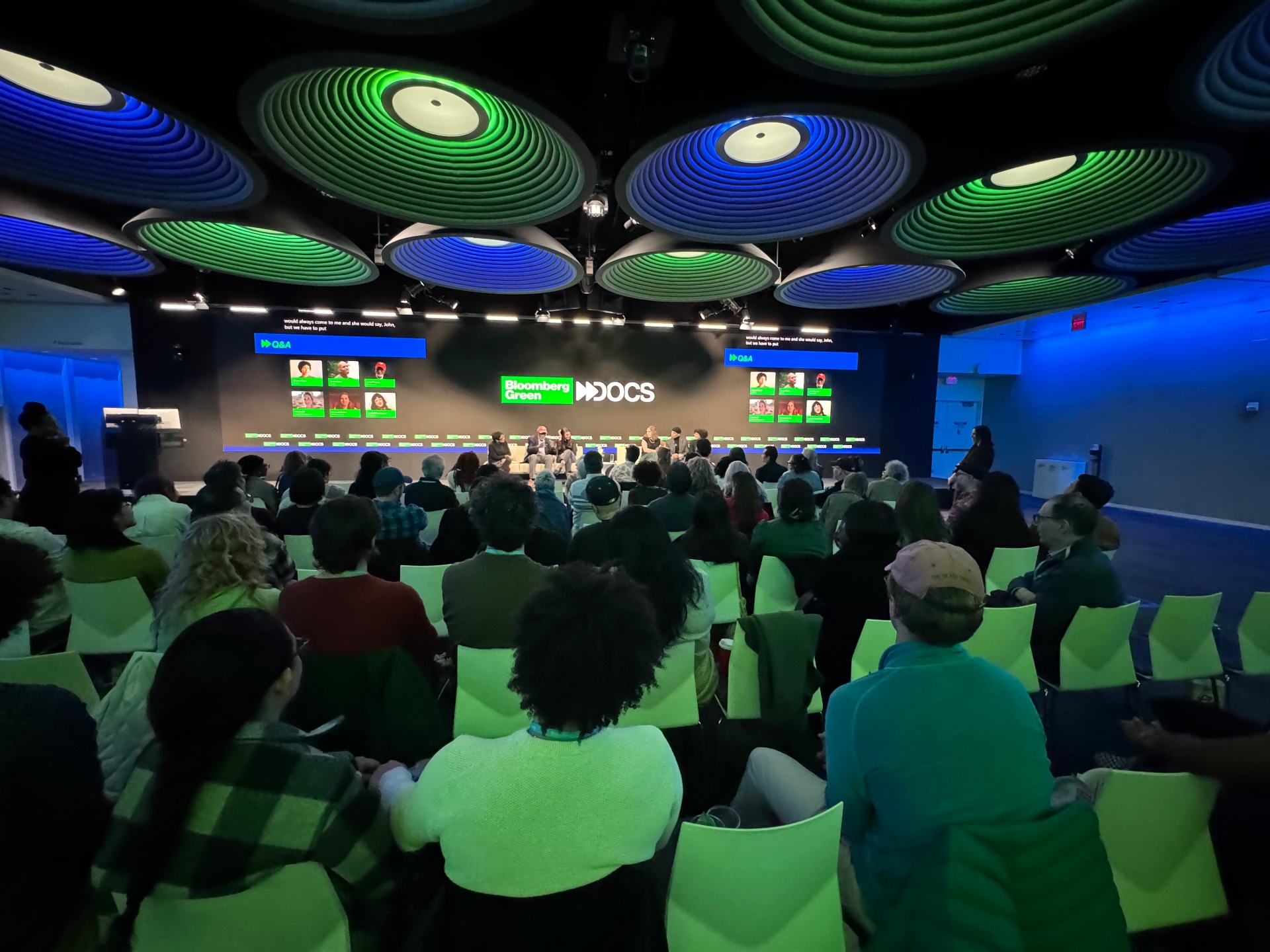 Audience seated for a Bloomberg Green Docs Q&A with on-screen title card and panelist headshots.